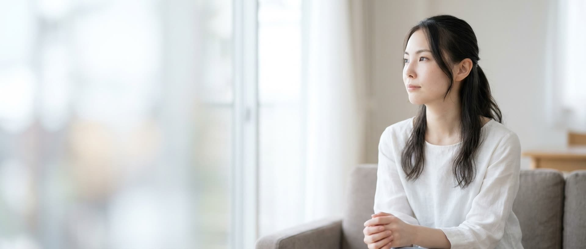 明るいリビングのソファで手を組み、窓のほうを見て静かに考える女性